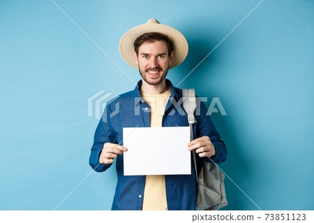 Happy tourist with backpack showing empty piece of paper, smiling at camera, standing on blue background. Concept of summer holiday and vacation Happy tourist with backpack showing empty piece of paper, smiling at camera, standing on blue background. Concept of summer holiday and vacation 73851123