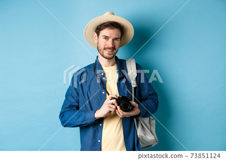 Handsome guy going on summer vacation, backpacking on holiday. Tourist with straw hat and camera smiling happy, standing on blue background 73851124