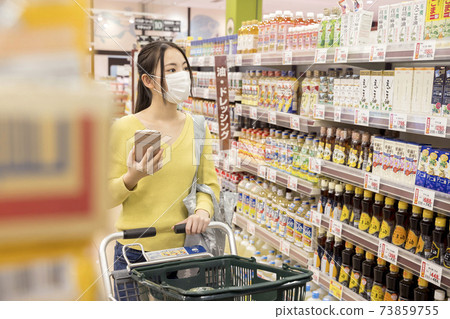A female shopper in a mask looking for a product while looking at a smartphone at a supermarket Shopping scene A female shopper in a mask looking for a product while looking at a smartphone at a supermarket Shopping scene 73859755