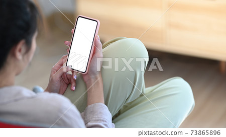 Rear view of young female sitting on couch in living room and using mobile phone. 73865896
