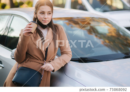 Beautiful young woman in black face mask stand by the car and drink coffee. Happy young woman have a break. Female pulled off mask for drinking cappucino outdoors Beautiful young woman in black face mask stand by the car and drink coffee. Happy young woman have a break. Female pulled off mask for drinking cappucino outdoors 73868092