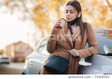 Beautiful young woman in black face mask stand by the car and drink coffee. Happy young woman have a break. Female pulled off mask for drinking cappucino outdoors 73868093