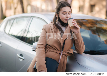 Beautiful young woman in black face mask stand by the car and drink coffee. Happy young woman have a break. Female pulled off mask for drinking cappucino outdoors Beautiful young woman in black face mask stand by the car and drink coffee. Happy young woman have a break. Female pulled off mask for drinking cappucino outdoors 73868094