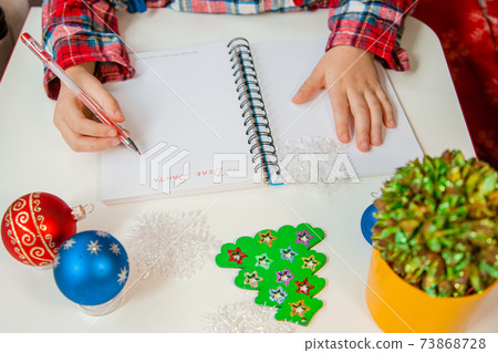 Dear Santa letter, Christmas card. A child holding a pen writes on a white sheet on a wooden background with New Year's decor. Dear Santa letter, Christmas card. A child holding a pen writes on a white sheet on a wooden background with New Year's decor. 73868728