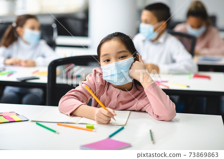 Asian school girl in mask sitting at desk in classroom 73869863