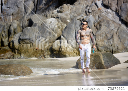 Athletic tattooed guy walking at the tropical beach Athletic tattooed guy walking at the tropical beach 73879621