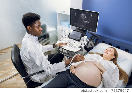 Young pretty pregnant woman getting ultrasound scan from doctor. Professional confident African man doctor obstetrician, checking baby in the belly with ultrasound transducer scanner. Young pretty pregnant woman getting ultrasound scan from doctor. Professional confident African man doctor obstetrician, checking baby in the belly with ultrasound transducer scanner. 73879947