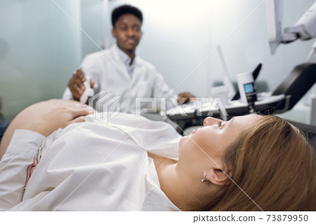 Cropped close up of young pregnant woman, visiting her male African doctor sonographer, having ultrasound scanning at the medical clinic. Ultrasound equipment, technology concept Cropped close up of young pregnant woman, visiting her male African doctor sonographer, having ultrasound scanning at the medical clinic. Ultrasound equipment, technology concept 73879950