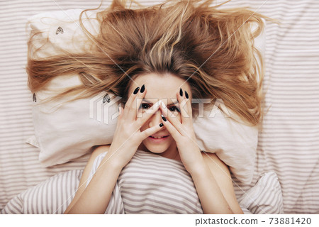 blonde woman lying in bed under a blanket holding her hands in front of her face and smiling blonde woman lying in bed under a blanket holding her hands in front of her face and smiling 73881420