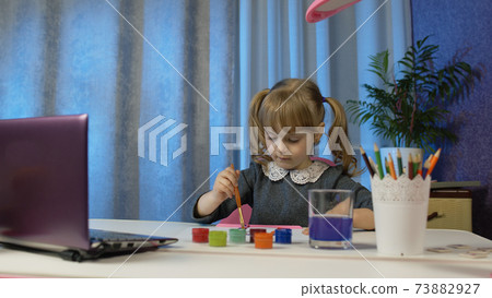 Child girl pupil enthusiastically drawing with brushes and paints, using laptop computer at home 73882927