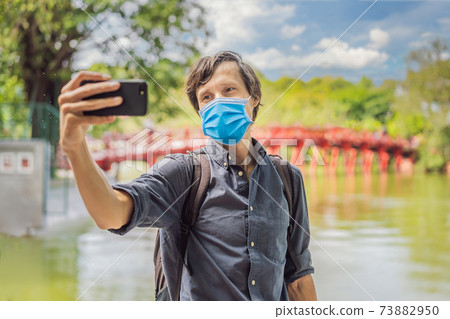 Caucasian man traveler wearing a medical mask during COVID-19 coronavirus on background of Red Bridge in public park garden with trees and reflection in the middle of Hoan Kiem Lake in Downtown Hanoi 73882950