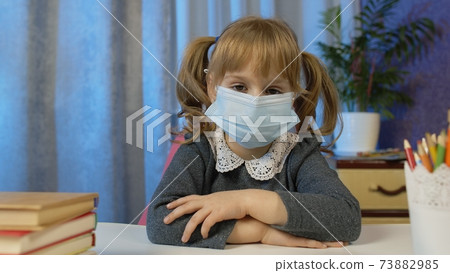 Sad child girl pupil in medical mask doing school homework at home, video call by webcam on internet Sad child girl pupil in medical mask doing school homework at home, video call by webcam on internet 73882985