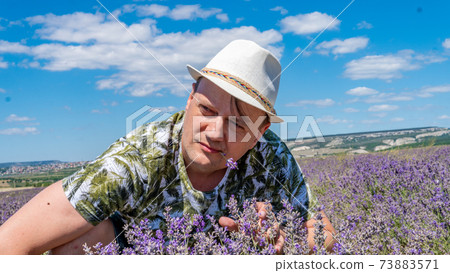 Smiling guy in lilac field looks seriously at the flowers sitting in a cap against the blue sky 73883571