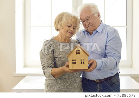 Smiling senior couple looking at a wooden model of a house being held in their hands. 73883572