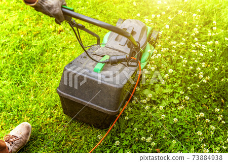 Man cutting green grass with lawn mower in backyard. Gardening country lifestyle background. Beautiful view on fresh green grass lawn in sunlight, garden landscape in spring or summer season. Man cutting green grass with lawn mower in backyard. Gardening country lifestyle background. Beautiful view on fresh green grass lawn in sunlight, garden landscape in spring or summer season. 73884938