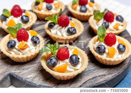 Fruit and berry tartlets dessert assorted on wooden tray. Closeup of delicious pastry sweets pies colorful cakes with fresh natural raspberry blueberry and cheese cream. French bakery catering. 73885000