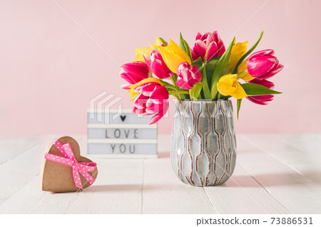 Recycled Heart shape gift box, spring bouquet of pink and yellow tulip flowers in a vase and Lightbox with love you message on the white wooden table and pink background. Greeting card mockup. Recycled Heart shape gift box, spring bouquet of pink and yellow tulip flowers in a vase and Lightbox with love you message on the white wooden table and pink background. Greeting card mockup. 73886531