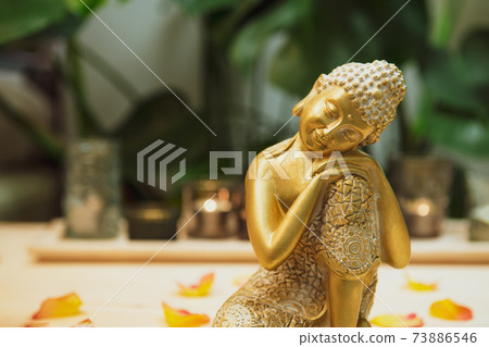Golden bronze Buddha decorative statuette with flower petals on the wooden table with burnig candles, monstera plant background. Meditation ritual, Spirituality, buddhism and religion. Selective focus 73886546