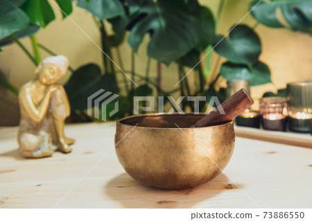 Tibetan singing bowl with special stick, burning candles and Golden Buddha decorative statuette on the wooden table with green plants background. Meditation and Relax. Exotic massage. Selective focus Tibetan singing bowl with special stick, burning candles and Golden Buddha decorative statuette on the wooden table with green plants background. Meditation and Relax. Exotic massage. Selective focus 73886550