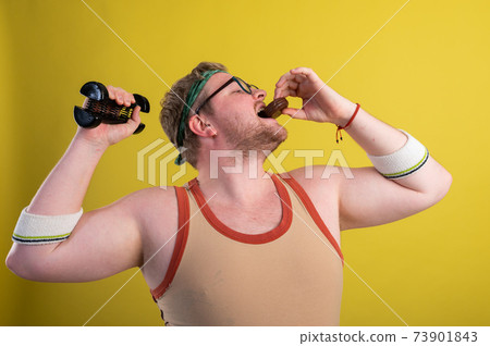 funny fat man in sports clothes with dumbbells in his hands eats chocolate cookies funny fat man in sports clothes with dumbbells in his hands eats chocolate cookies 73901843