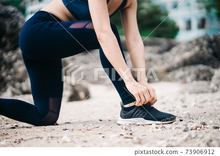 Asian fitness runner body closeup doing warm-up routine on beach before running, stretching leg muscles with standing single knee to chest stretch. Female athlete preparing legs for cardio workout. Asian fitness runner body closeup doing warm-up routine on beach before running, stretching leg muscles with standing single knee to chest stretch. Female athlete preparing legs for cardio workout. 73906912