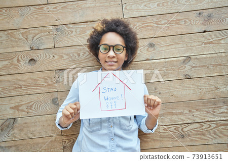 Mixed race teen girl holding drawing picture with words Stay Home while standing over wooden wall background. Social media campaign for coronavirus prevention 73913651