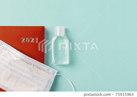 Medical face mask on a 2021 year red leather diary and hand sanitizer on a turquoise background.  73915963