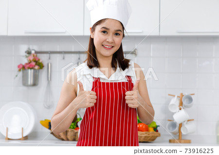 Portrait of happy Asian woman chef in red apron standing in kitchen. 73916225