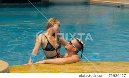 The loving couple hugs and kisses, drinking blue cocktail alcohol liquor in swimming pool at hotel outdoor. Portrait of caucasian man and woman. The loving couple hugs and kisses, drinking blue cocktail alcohol liquor in swimming pool at hotel outdoor. Portrait of caucasian man and woman. 73920417