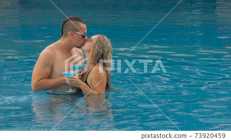 The loving couple hugs and kisses, drinking blue cocktail alcohol liquor in swimming pool at hotel outdoor. Portrait of caucasian man and woman. The loving couple hugs and kisses, drinking blue cocktail alcohol liquor in swimming pool at hotel outdoor. Portrait of caucasian man and woman. 73920459