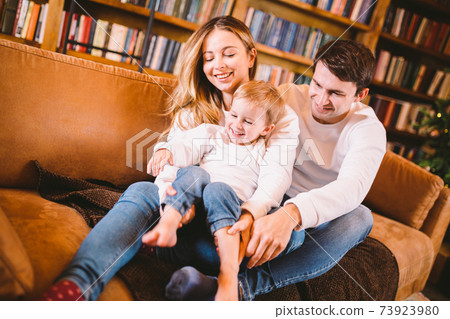 Lovely family with baby son dressed in the same clothes have fun on sofa at home during Christmas. Family Christmas Concept. Happy parents with cute child In a living room on new year holidays 73923980
