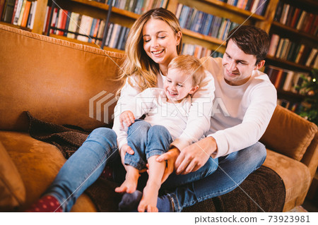 Christmas family portrait. Young Caucasian family in identical clothes having fun on sofa in living room with large library of books and Christmas tree in evening. Happy New Year and Merry Christmas Christmas family portrait. Young Caucasian family in identical clothes having fun on sofa in living room with large library of books and Christmas tree in evening. Happy New Year and Merry Christmas 73923981