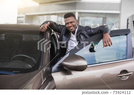 Close up portrait of young handsome African guy, wearing black leather jacket, posing at petrol station to camera with smile, while leaning on the open door of his lexury car Close up portrait of young handsome African guy, wearing black leather jacket, posing at petrol station to camera with smile, while leaning on the open door of his lexury car 73924112