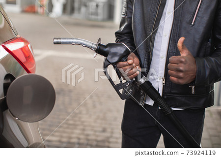 Close up cropped image of unrecognizable black skinned man in black casual outfit, standing outdoors at the petrol station and holding nozzle of fuel gun in hands and showing his thumb up Close up cropped image of unrecognizable black skinned man in black casual outfit, standing outdoors at the petrol station and holding nozzle of fuel gun in hands and showing his thumb up 73924119