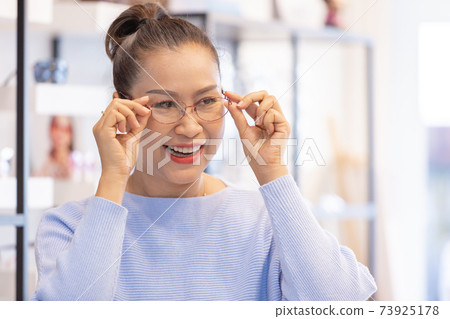Middle aged asian woman choosing spectacles glasses in optician store. 73925178