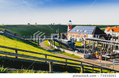 Westkapelle, The Netherlands, August 2019. The built-up area, below sea level, is protected by an artificial hill that surrounds it. On the top of the hill a red road. Westkapelle, The Netherlands, August 2019. The built-up area, below sea level, is protected by an artificial hill that surrounds it. On the top of the hill a red road. 73938091