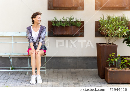 Young adult beautiful caucasian woman sitting on metal vintage bench chair against wall with many mounted ruty plant pot on city street. Attractive female person waiting meeting outdoors in garden 73944426