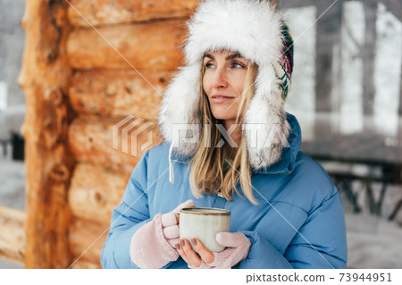 Portrait of a caucasian woman in a winter hat with earflaps with a mug of hot drink. Portrait of a caucasian woman in a winter hat with earflaps with a mug of hot drink. 73944951