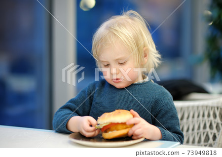 Cute preschooler boy eating large hamburger at fast food restaurant 73949818