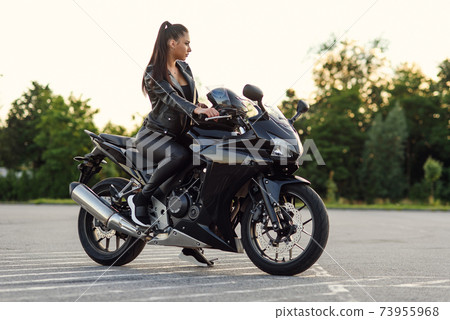 Stylish beautiful girl in black leather jacket and pants on outdoors parking sits on sports motorcycle and holds protective helmet. Stylish beautiful girl in black leather jacket and pants on outdoors parking sits on sports motorcycle and holds protective helmet. 73955968