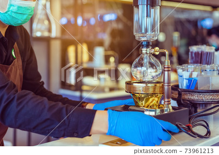 Syphon (Siphon) coffee maker brews drink using two chambers where vapor pressure and vacuum produce coffee with barista wear surgical mask and gloves prepare to brewing in cafe. Selective focus. 73961213