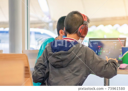 Back view of Asian boy wearing protective face mask and headphones while working with a friend on laptop during corona virus (Covid-19) and flu outbreak in food court at the mall.New normal lifestyle. Back view of Asian boy wearing protective face mask and headphones while working with a friend on laptop during corona virus (Covid-19) and flu outbreak in food court at the mall.New normal lifestyle. 73961580