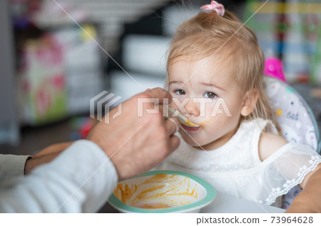A caring man with a beard feeds his little daughter. A little girl sits at a children's dining table and dines with her dad. Happy fatherhood. A caring man with a beard feeds his little daughter. A little girl sits at a children's dining table and dines with her dad. Happy fatherhood. 73964628