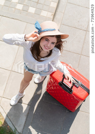 Top view on beautiful young caucasian woman in summer hat posing with red suitcase outdoors. Happy smiling girl goes on vacation with a large luggage bag. Charming lady in shorts on a hot sunny day. 73964929