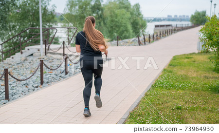 An obese young woman jogging outdoors. Fat beautiful smiling girl in a black tracksuit is engaged in fitness for weight loss on the waterfront. A woman runs on a summer day. 73964930