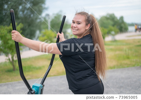 Beautiful smiling overweight young woman does fitness on an ellipsoid simulator outdoors. Fat girl is training on the sports ground for weight loss. Sport outside on a warm summer day. 73965005