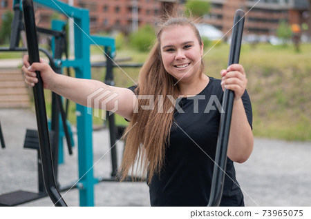 Beautiful smiling overweight young woman does fitness on an ellipsoid simulator outdoors. Fat girl is training on the sports ground for weight loss. Sport outside on a warm summer day. 73965075
