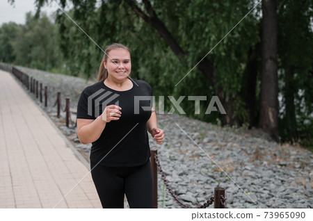 An obese young woman jogging outdoors. Fat beautiful smiling girl in a black tracksuit is engaged in fitness for weight loss on the waterfront. A woman runs on a summer day. 73965090