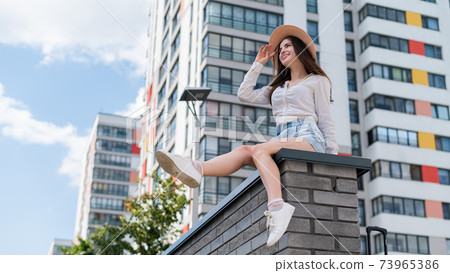 Beautiful young woman in shorts and a hat posing while sitting on a stone bench. A full-length portrait of a Caucasian girl on a hot summer day. Beautiful young woman in shorts and a hat posing while sitting on a stone bench. A full-length portrait of a Caucasian girl on a hot summer day. 73965386