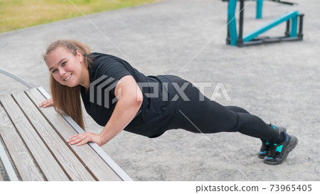 Fat young woman push up from the bench outdoors. A chubby smiling girl warms up before jogging. Sports for weight loss. Fat young woman push up from the bench outdoors. A chubby smiling girl warms up before jogging. Sports for weight loss. 73965405